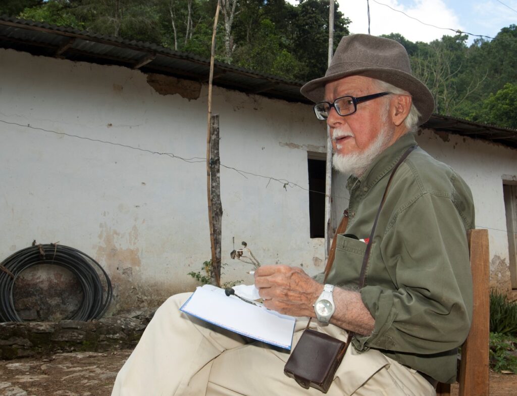 Gastón Guzmán sitting in a chair with a notebook on his lap and a pair of glasses in his hand.