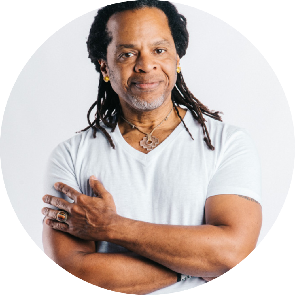 Tony Moss - a black man with dreadlocks wearing a white, v-neck t-shirt poses with his hands crossed in front of him. He has a slight smile on his face. 