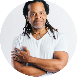 Tony Moss - a black man with dreadlocks wearing a white, v-neck t-shirt poses with his hands crossed in front of him. He has a slight smile on his face.