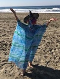 Redwing Keyssar standing on a beach wearing a witch hat, arms spread wide in the air.