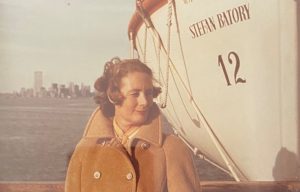 Jane Osmond, a middle-aged white woman, stands in front of a ship called the "Stefan Batory" in the New York City Harbour. Jane is wearing an orange-tan peacoat and is gazing to the right.