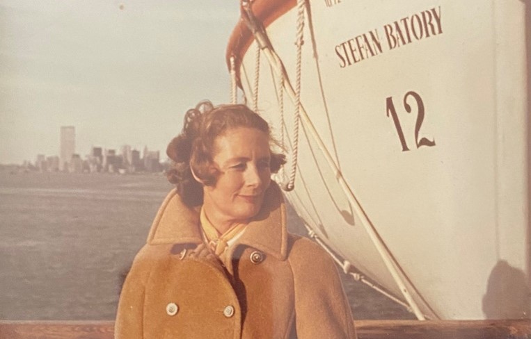 Jane Osmond, a middle-aged white woman, stands in front of a ship called the "Stefan Batory" in the New York City Harbour. Jane is wearing an orange-tan peacoat and is gazing to the right.