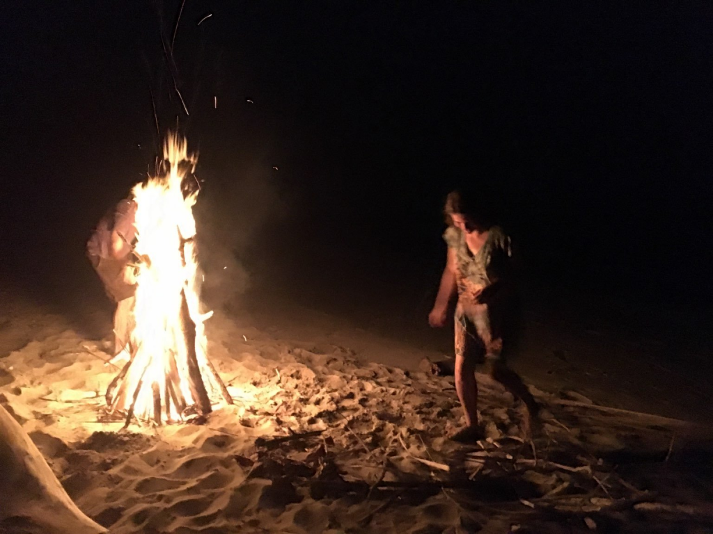 An psychedelic psychotherapist dances around a bonfire on a beach.