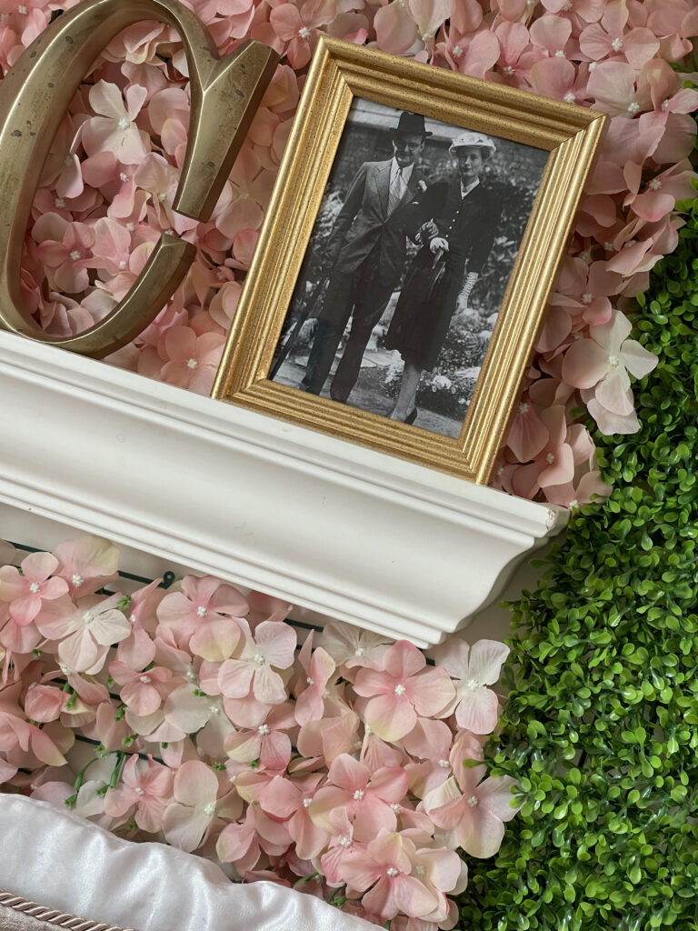 A picture frame with a photo of Jane and Humphry Osmond sits on a white shelf with a letter "C" figure and a background of pink flowers.