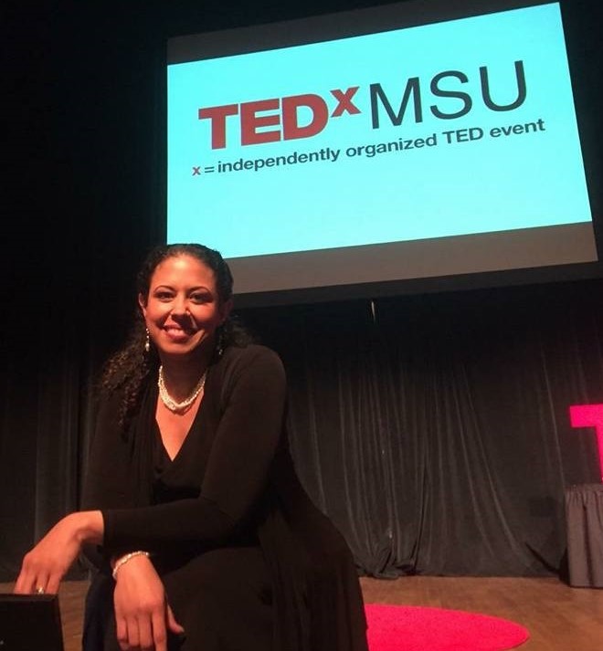 NiCole Buchanan, a black woman with long hair wearing a black outfit, sits in front of the Ted x MSU sign.