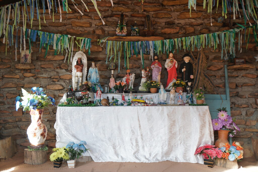 a table with a white table cloth with different idols lined up on it. Vases with flowers are on the floor beside the table. The wall in the background is made of stone. Streamers hang from the ceiling.