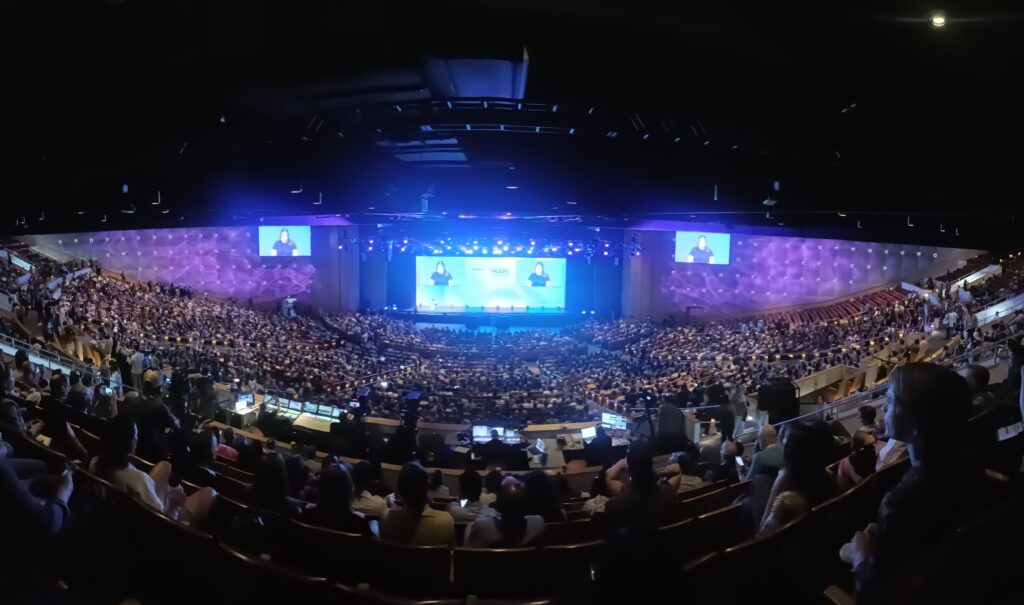 A vision from the back of a large theater that shows the opening talks at Psychedelic Science 2023 in Denver Colorado. The stage is lit in blue and pink light. The audience is full. 